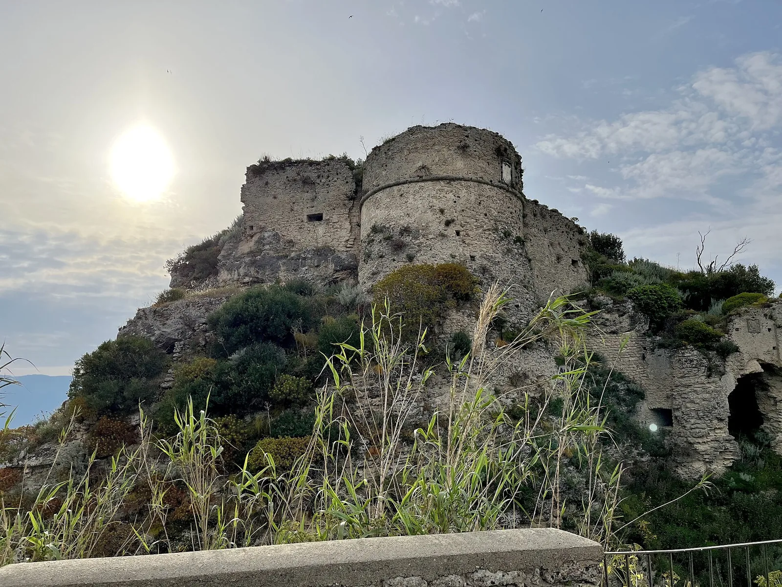 Ruins of Gerace castle above shrubs, under bright sky.